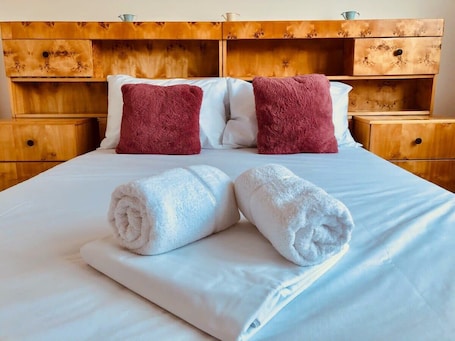 A neatly made bed with two rolled white towels and two pillows, one white and one maroon, in a room with wooden headboard and nightstands.