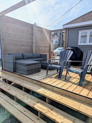 A wooden deck with a sofa and chairs, a grill, and a house in the background.