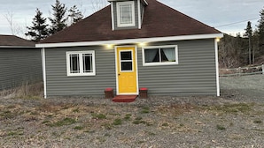 Una casa de un piso con puerta principal amarilla, dos ventanas y un porche pequeño.