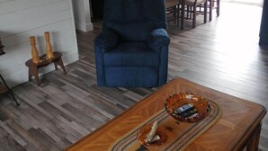 A living room with a blue armchair, wooden coffee table with remote controls, a dining area with chairs, and a kitchen area with cabinets and a clock. - Hatchet Harbour Vacation Home (Herring Neck)