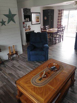 A living room with a blue armchair, wooden coffee table with remote controls, a dining area with chairs, and a kitchen area with cabinets and a clock.