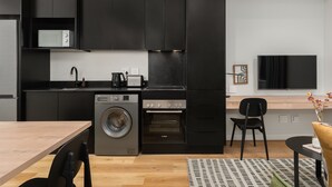 A modern kitchen with black cabinets, a microwave, a washing machine, and an oven. There is a dining table with chairs and a TV on the wall. - Cozy Studio by Table Mountain Explore Cape Town (Cape Town)