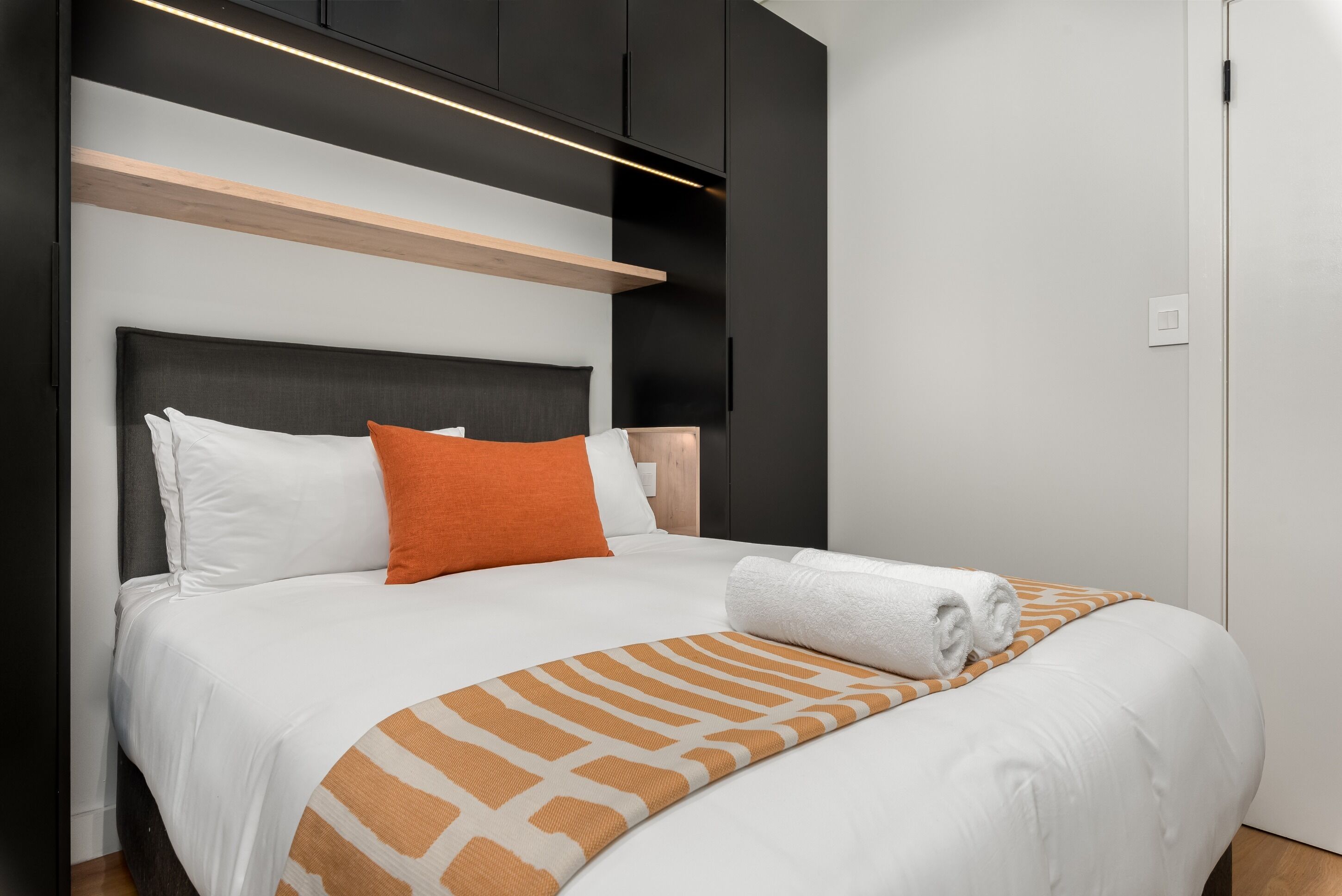 Un letto ben rifatto con biancheria da letto bianco e arancione, una testiera in legno e una mensola a muro sopra.