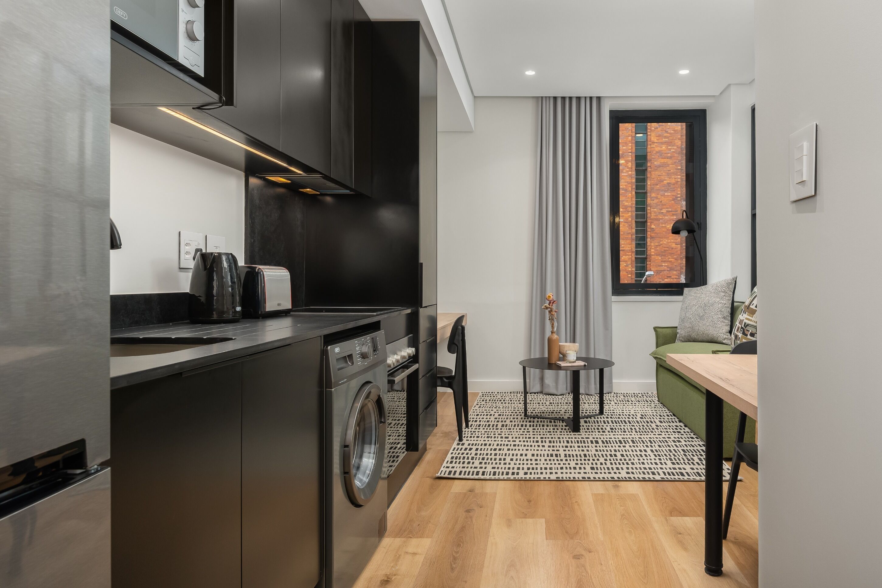 A compact kitchen with modern appliances, a dining area with a small table and chairs, and a view of a brick building through a window.