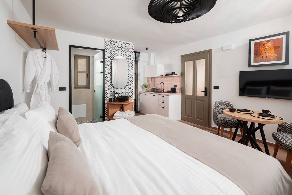 A modern hotel room with a large bed, a small kitchenette, a dining area, and a TV.
