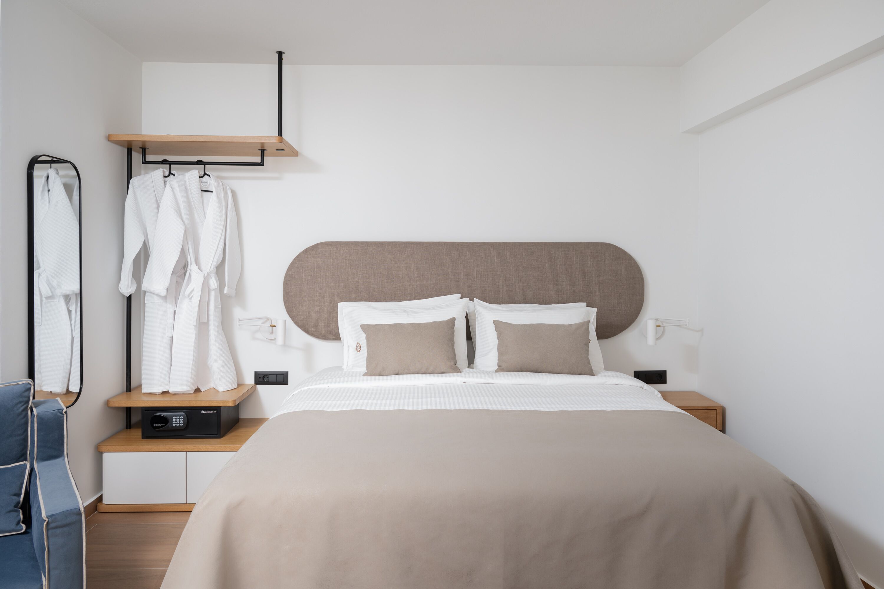 A modern hotel room with a large bed, a bedside table, a robe rack, and a wall-mounted mirror.