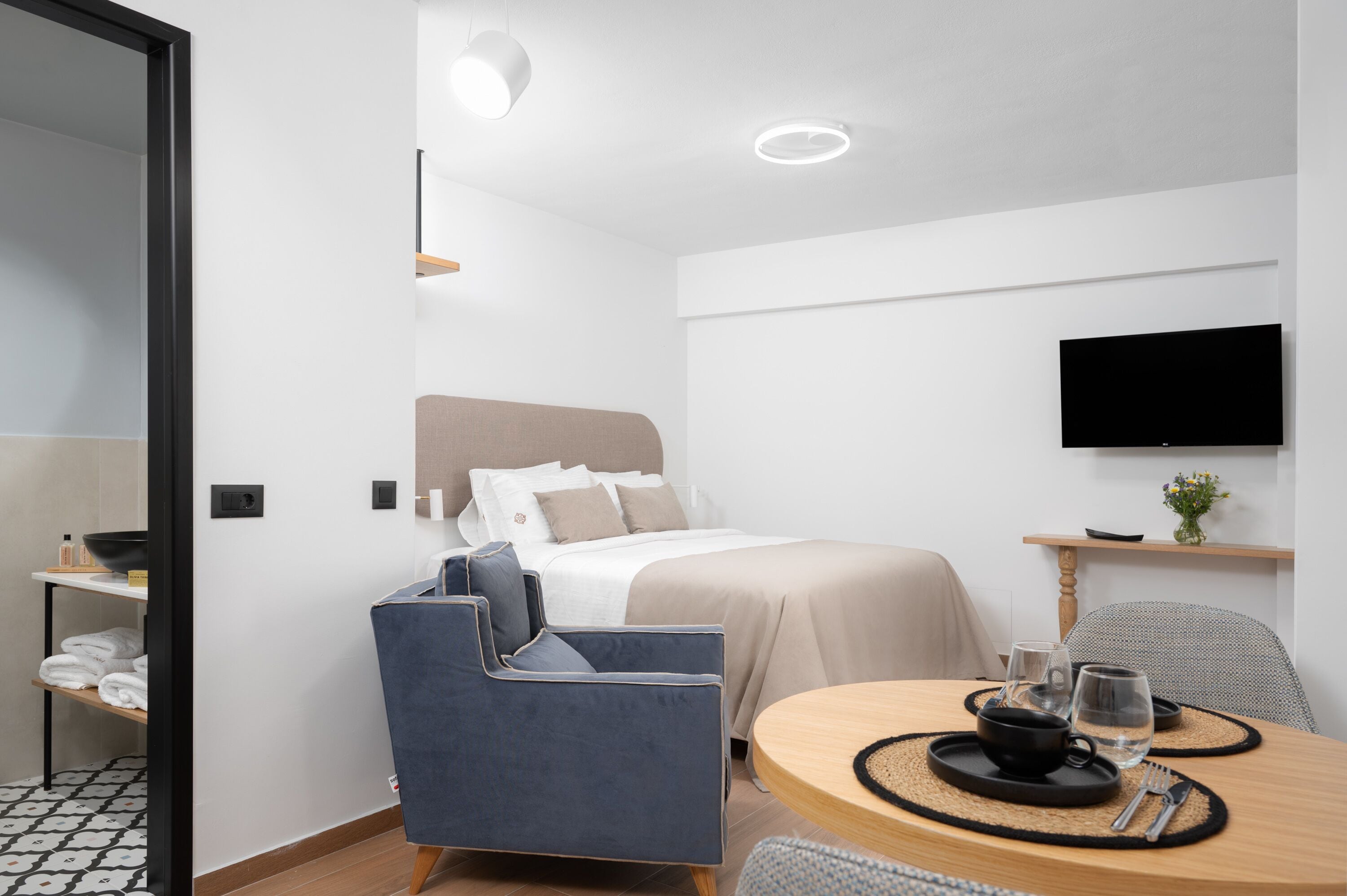 Et moderne hotelværelse med en seng, et siddeområde med et bord, et fjernsyn ophængt på væggen og en lille reol med en vase.