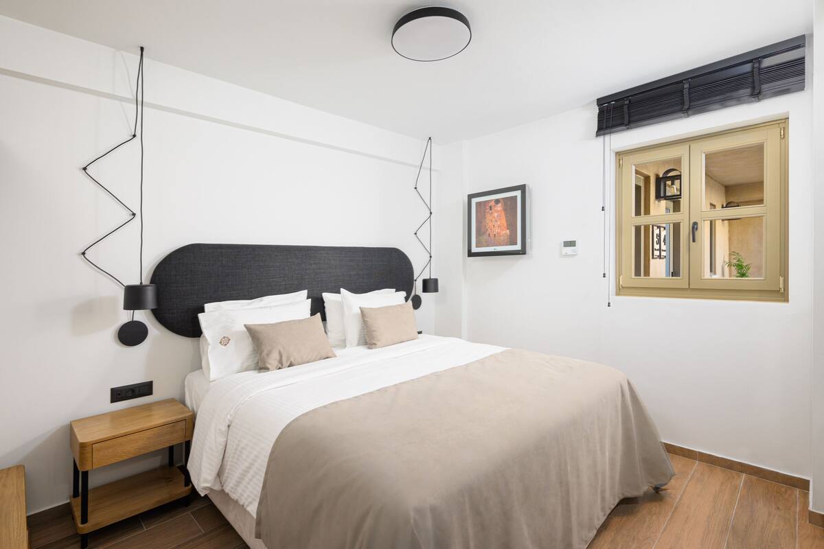 A modern bedroom with a large bed, a bedside table, a wall-mounted lamp, and a window with a view of the interior.