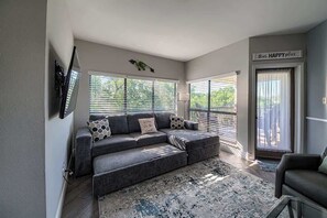 A living room with a grey sectional sofa, a glass coffee table, and a large window with blinds.