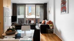 A modern living room with a sofa, a glass coffee table, and a TV. There is a large window with curtains, a red bus artwork on the wall, and a view of buildings outside. - The heart of the CBD - 2BR Modern Apt Wi-Fi (Adelaide)