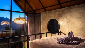 Un dormitorio con un ventanal grande, una cama con cubrecama de cuadros, un espejo redondo y una lámpara de techo tejida.