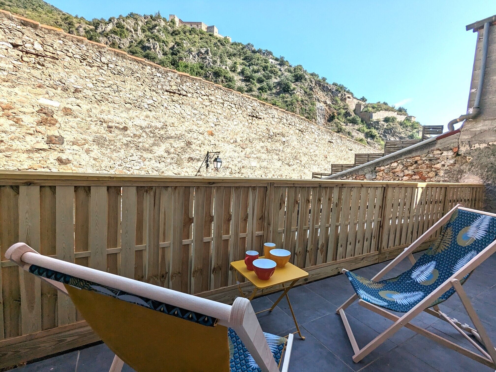 A wooden deck with two lounge chairs, a small table with red cups, and a stone wall.