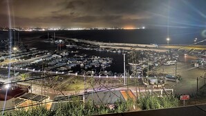 A marina at night with boats docked, a stormy sky, and city lights in the distance.