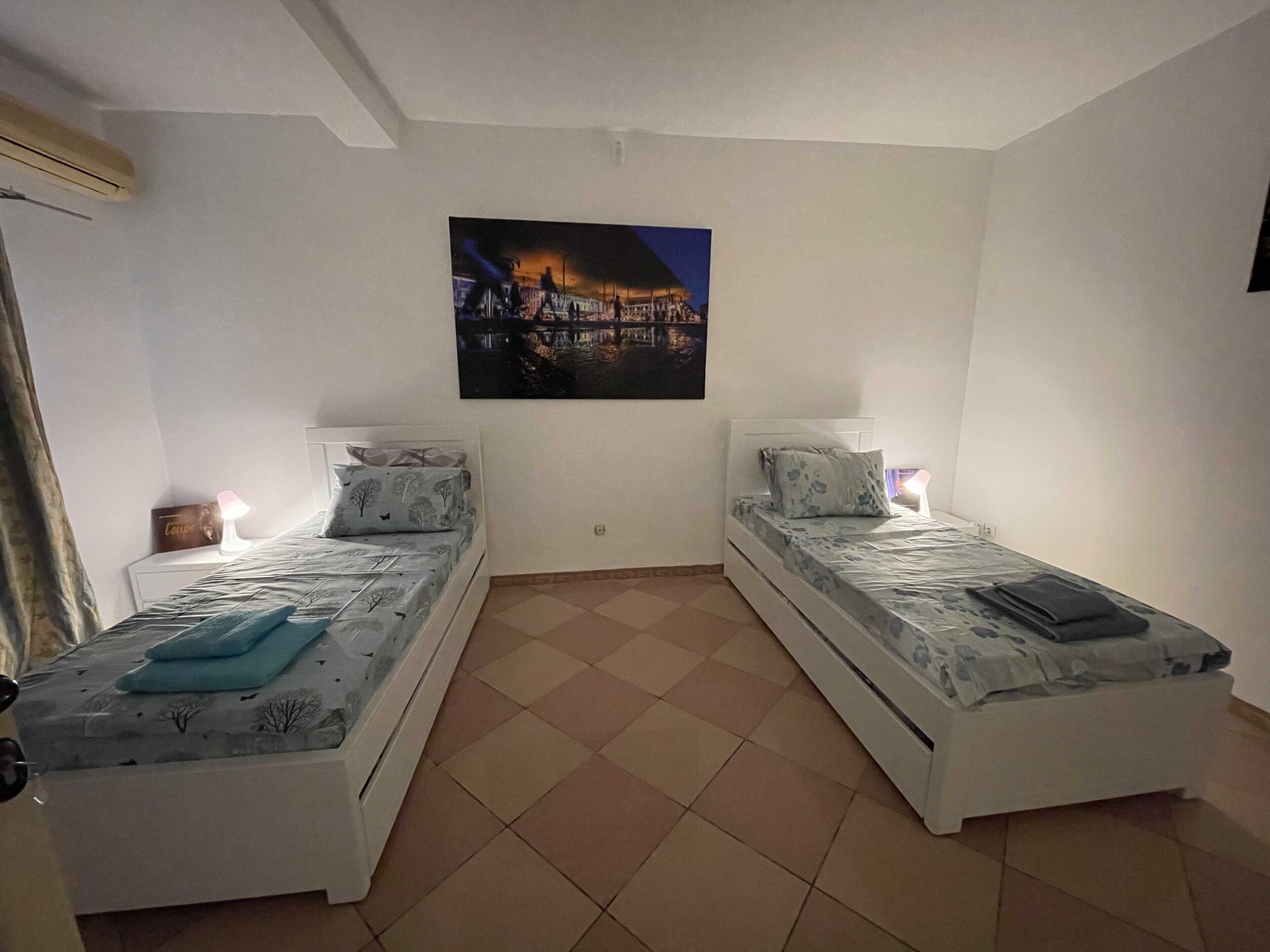 A room with two single beds, a nightstand with a lamp, a wall-mounted painting, and a tiled floor.