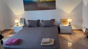 A bedroom with a large bed, two nightstands, a painting of a desert landscape, and a view of the sky.