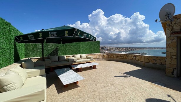 An outdoor seating area with a green umbrella, a stone wall, and a view of the sea and cityscape.