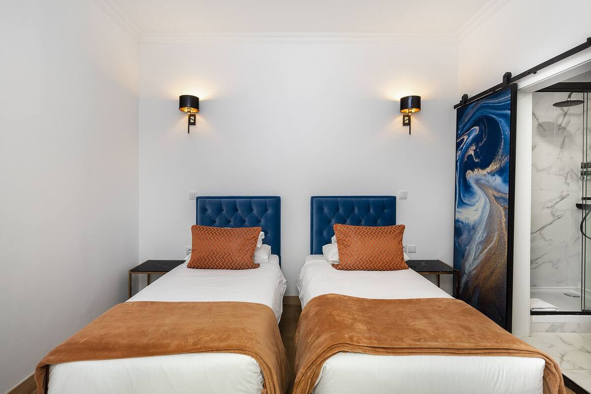 A hotel room with two beds, each with a blue headboard and brown bedding. There are wall-mounted lights and a large abstract painting on the wall.
