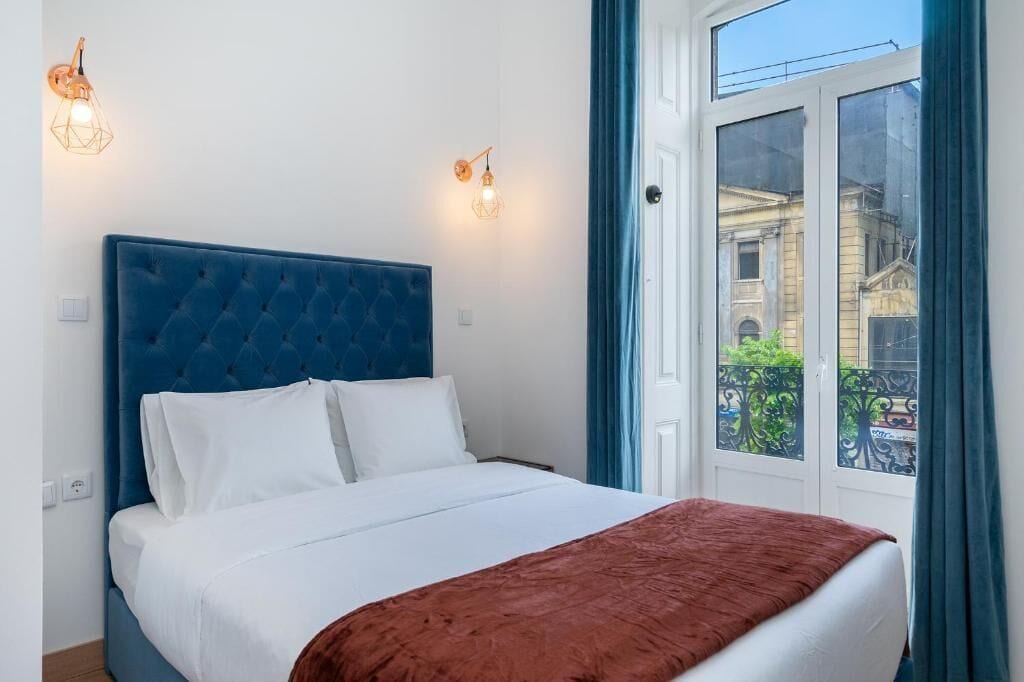 A bedroom with a blue headboard, white bedding, and a view of a building through a window.