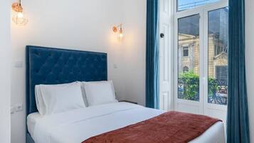 A bedroom with a blue headboard, white bedding, and a view of a building through a window.