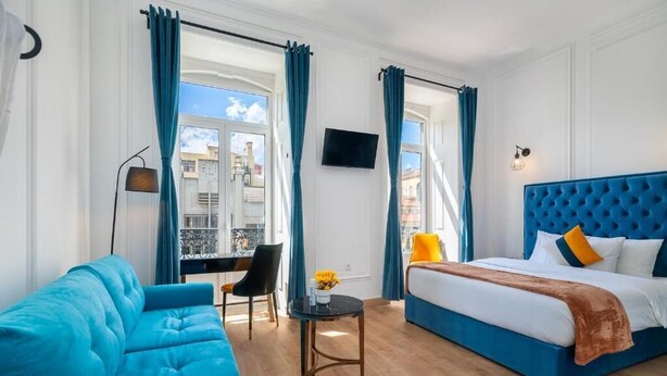 Una habitación de hotel con un sofá azul, una cama con cabecera azul, una mesita con un jarrón y un televisor en la pared.