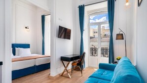 Una habitación moderna con un sofá azul, una cama con ropa blanca, un escritorio y una silla, un televisor montado en la pared, y un balcón con cortinas azules.
