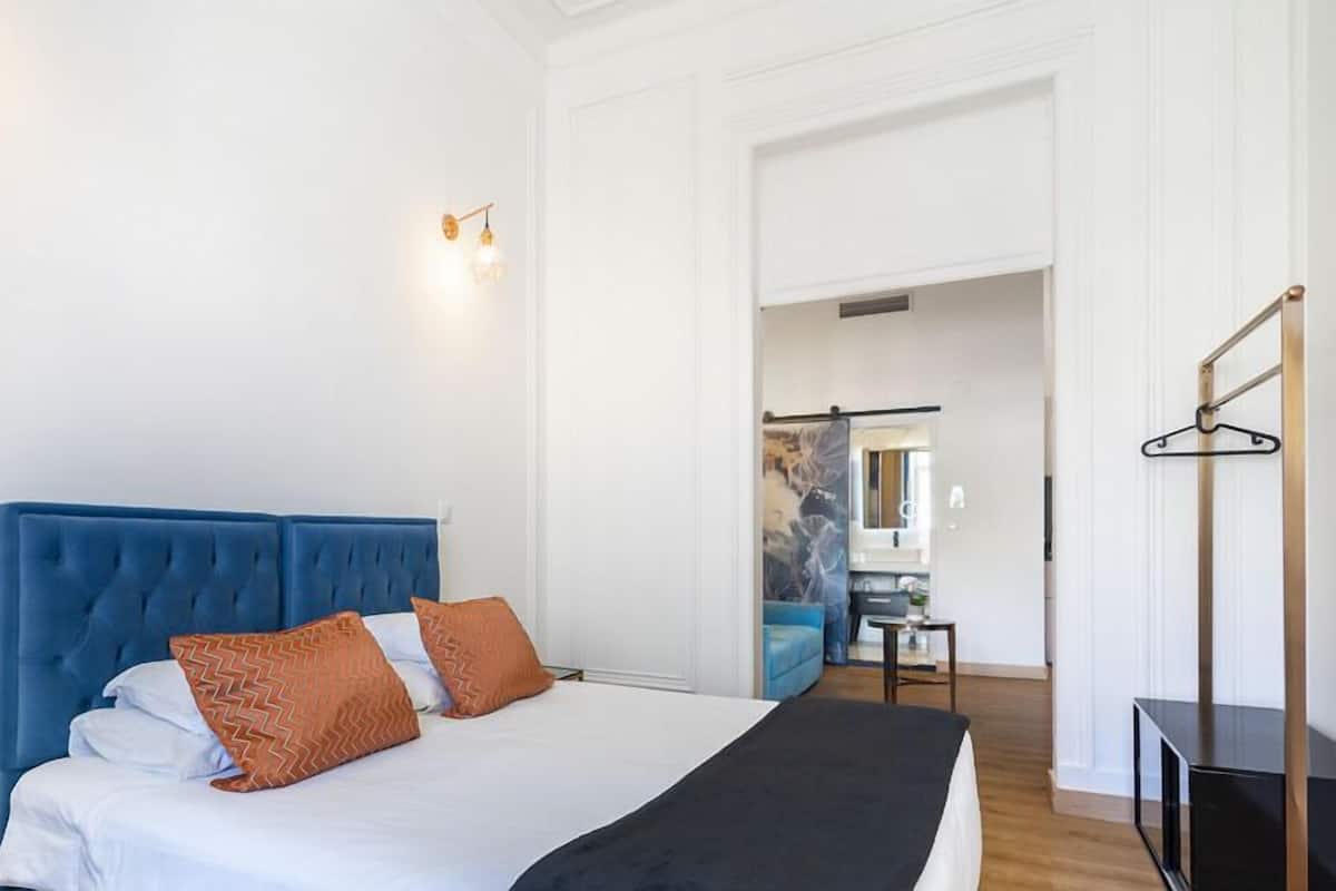 A hotel room with a large bed, a blue headboard, and a view of the bathroom and hallway.
