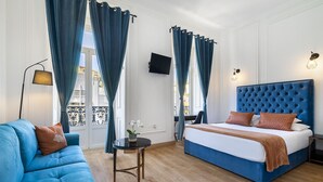 Habitación de hotel con un sofá azul, una cama con cabecera azul, una mesita redonda con una planta y un televisor montado en la pared.
