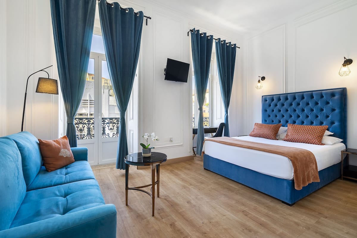 A hotel room with a blue sofa, a bed with a blue headboard, a small round table with a plant, and a TV mounted on the wall.