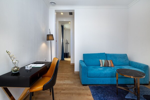 A modern living room with a blue sofa, a small round table, a black desk with a chair, and a lamp.