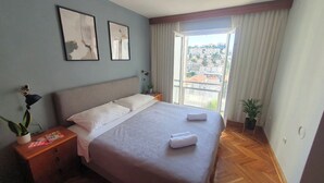 A neatly made bed with a patterned bedspread, two framed pictures on the wall, a nightstand with a lamp and a magazine, and a view of a cityscape through a window with curtains.