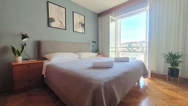 A bedroom with a large bed, a nightstand with a lamp, and a window with a view of buildings.