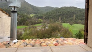 A view from a window overlooking a green valley with a chimney emitting smoke, indicating a fireplace or stove.