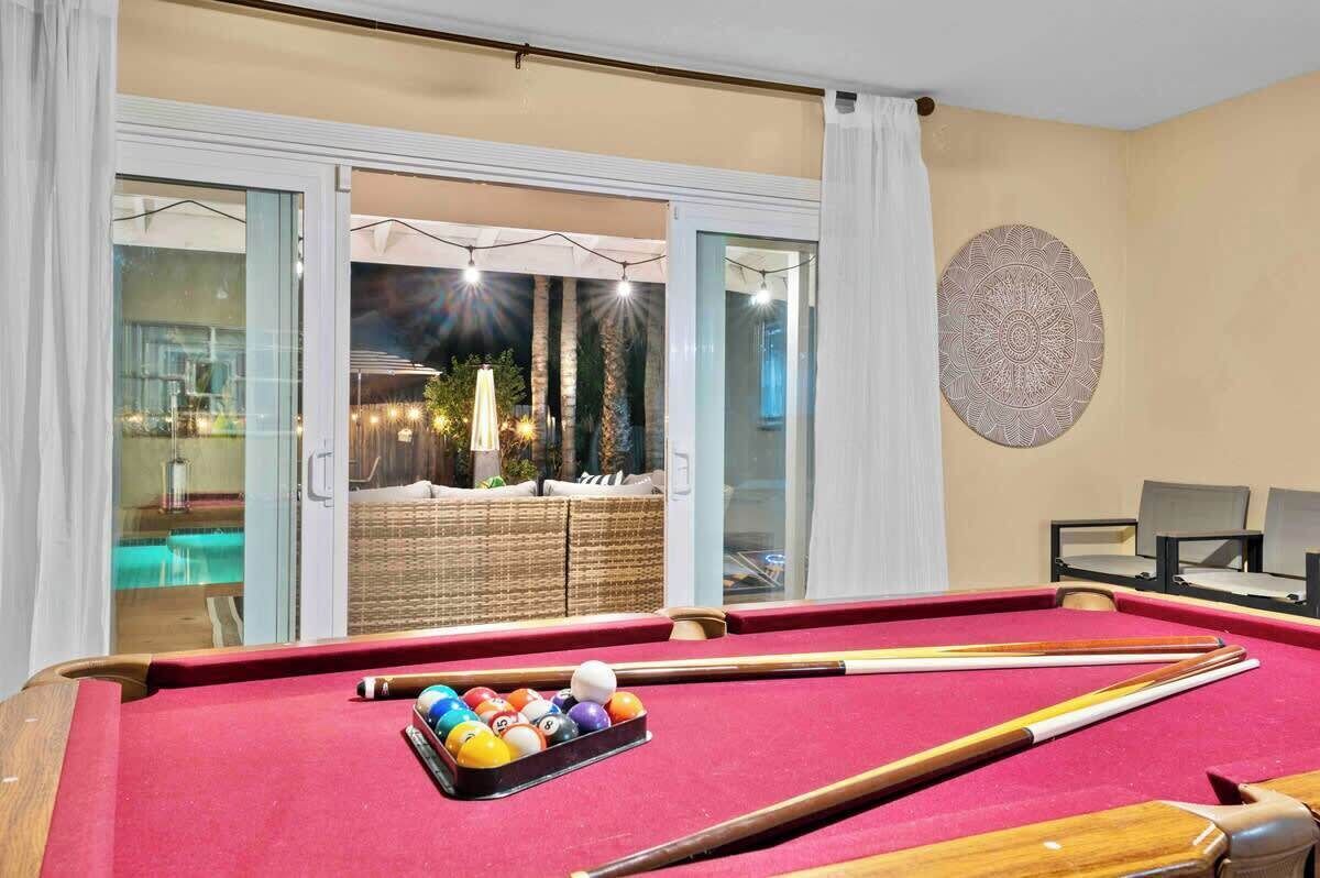 Une table de billard avec des billes et des queues, une chaise de table de billard, et une vue sur un espace de détente avec un canapé en osier et des éclairages.