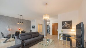 Una sala moderna con sofá, una mesa de centro de vidrio y una televisión. Se aprecia un comedor con sillas y una mesa, y una cocina al fondo.
