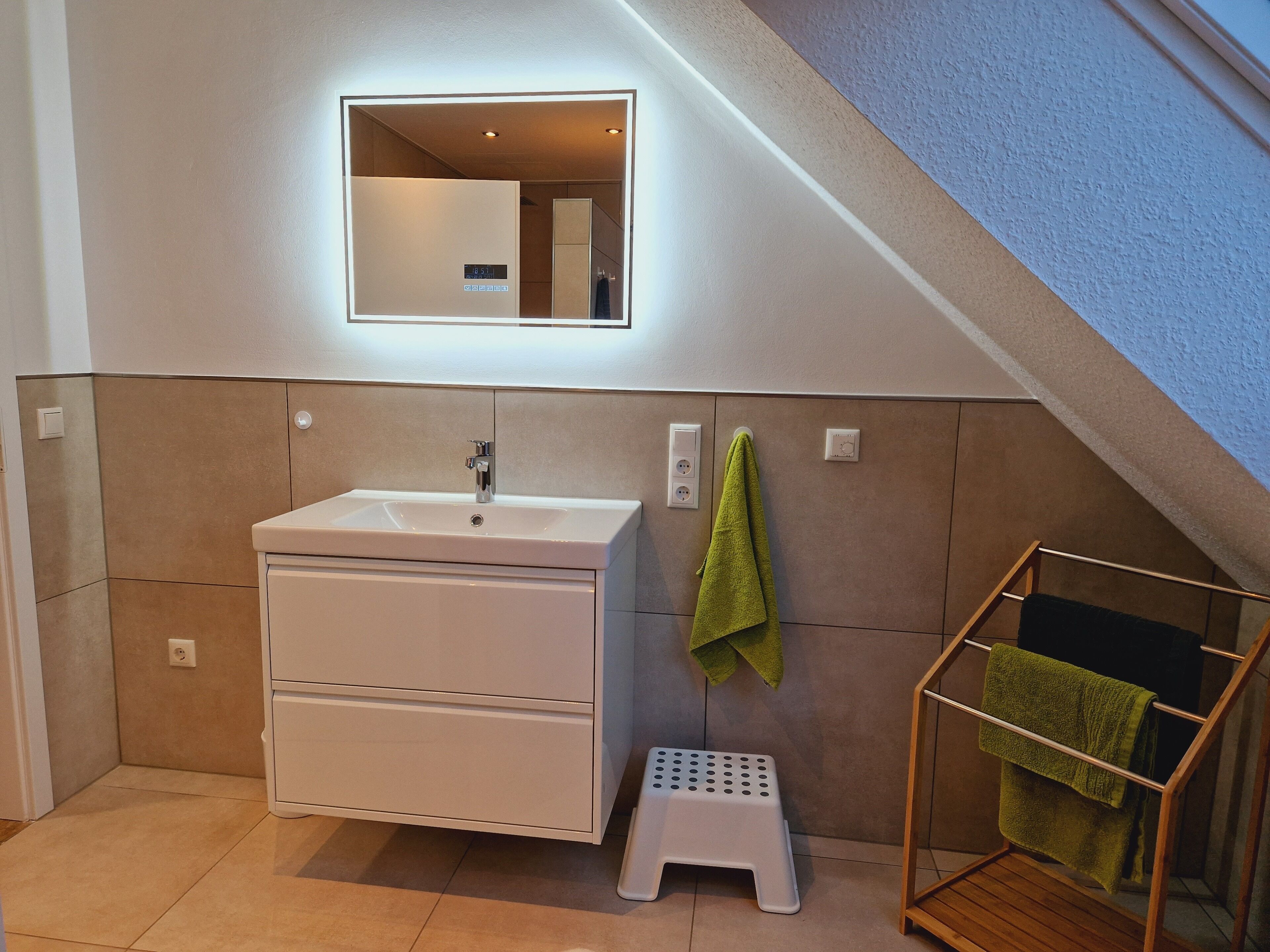 A modern bathroom with a white sink, mirror, and storage cabinet. A green towel hangs on a rack, and a small stool is placed near the sink.