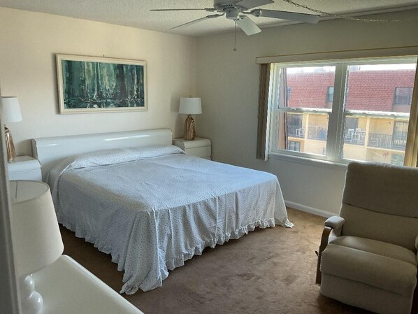Une chambre à coucher comprenant un lit, une chaise, une table de chevet, une lampe, un ventilateur de plafond et une fenêtre donnant sur des immeubles.