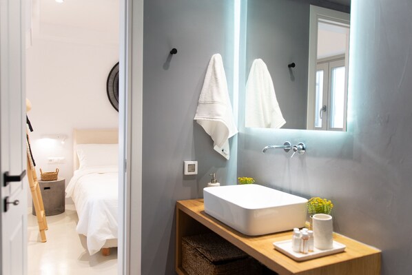 Un baño moderno con un lavamanos grande de color blanco, un espejo y un mueble de tocador de madera. Se observa una cama con ropa de cama blanca al fondo.