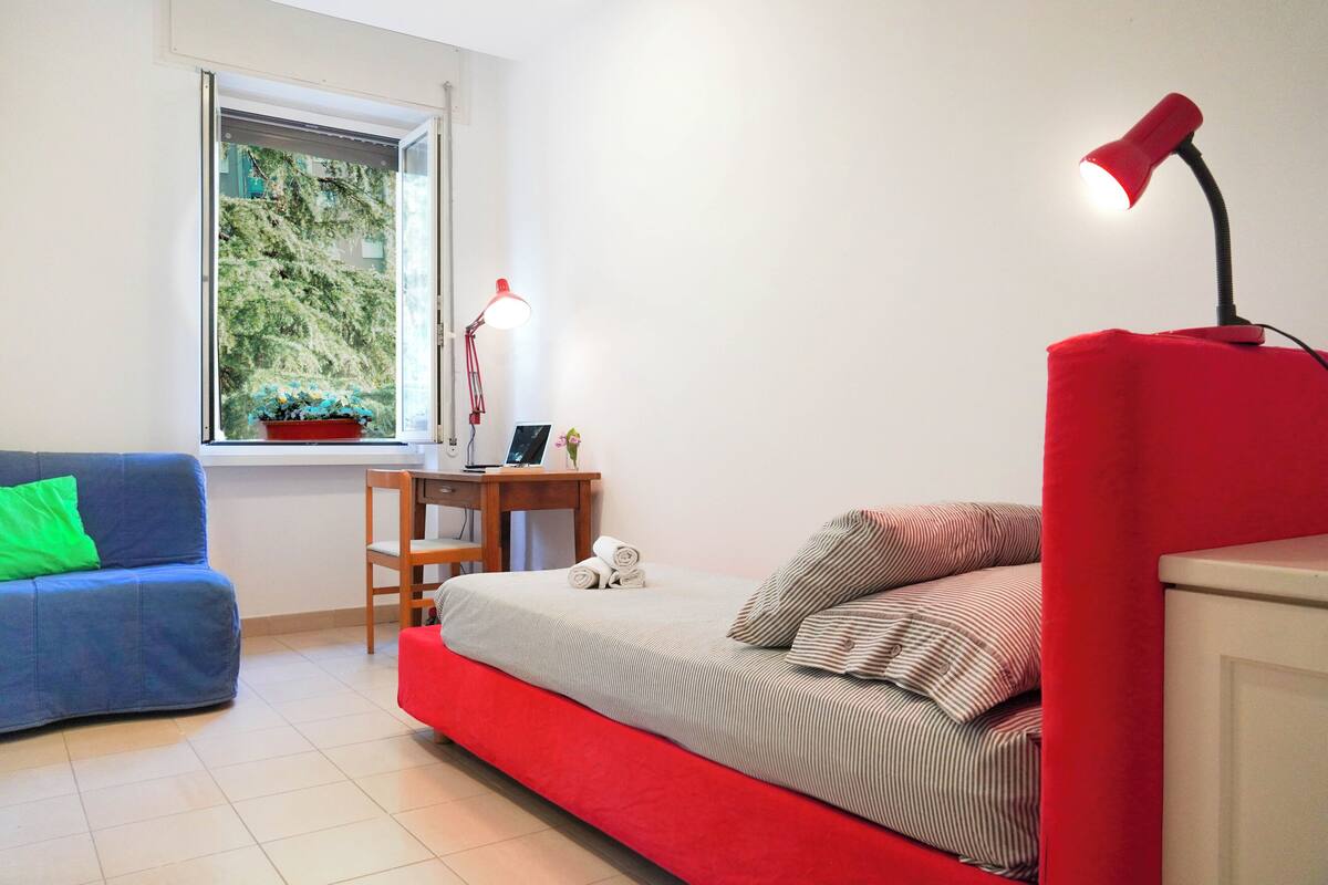 A compact bedroom with a bed, a desk, a chair, a window with a view of greenery, and a red lamp.