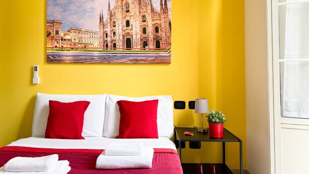 A hotel room with a bed, red and white pillows, a nightstand with a lamp, a small potted plant, and a framed picture of a cathedral on the wall. - CiaoMi - Hostel & Long Stay - Milano Niguarda & Bicocca  (Milan)