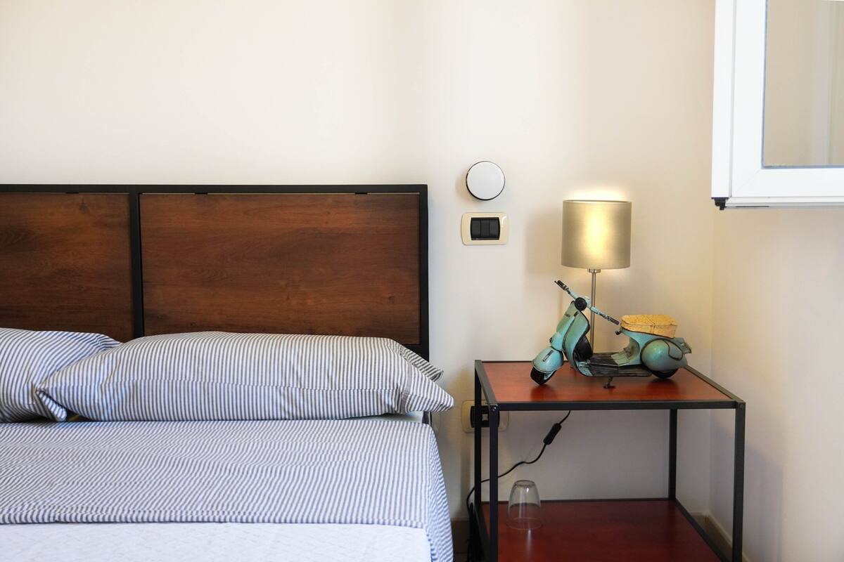 A neatly made bed with striped bedding, a wooden headboard, a bedside table with a lamp and a vintage scooter, and a wall-mounted light switch.