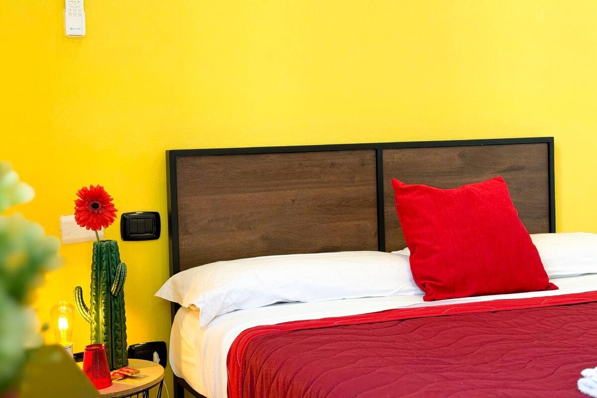 A bed with a red pillow and a red blanket, a wooden headboard, and a wall-mounted light switch.