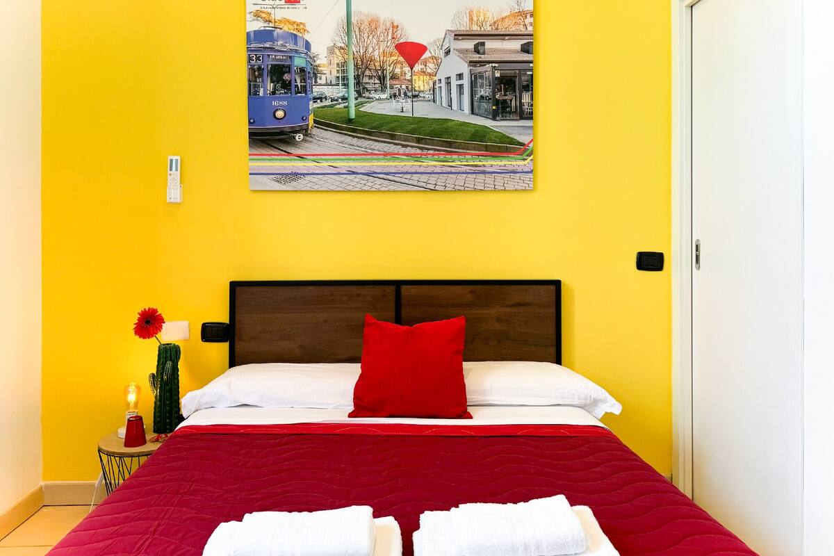 A hotel room with a bed, a red pillow, a nightstand with a lamp and a cactus, and a wall-mounted picture of a tram.