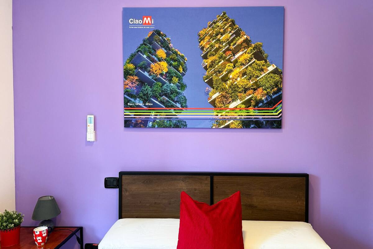 A bedroom with a bed, a red pillow, a nightstand with a lamp, a remote control, and a framed picture of a building with greenery on the wall.