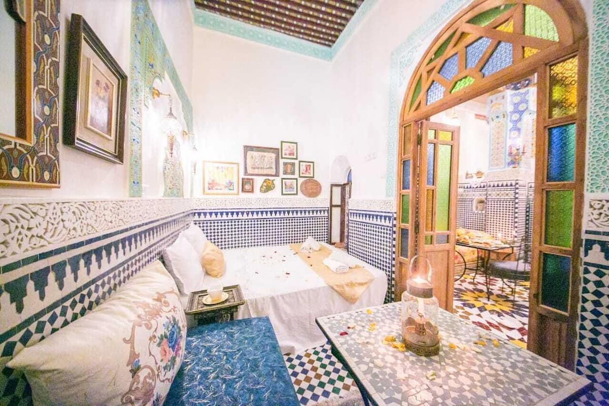A traditional Moroccan interior with a patterned tile floor, a sofa with a decorative pillow, a bed with a white bedspread, and a table with a candle.