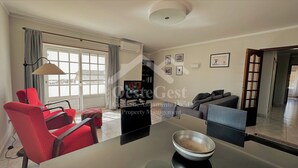 Uma sala de estar moderna com uma mesa de jantar, cadeiras vermelhas, um sofá cinzento e uma janela grande com cortinas.