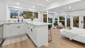 A modern kitchen with white cabinetry and a central island, a bedroom with a large bed and a view of the outdoors, and a dining area with a table and chairs.