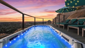 A hot tub with a city view at sunset, surrounded by lounge chairs and string lights.