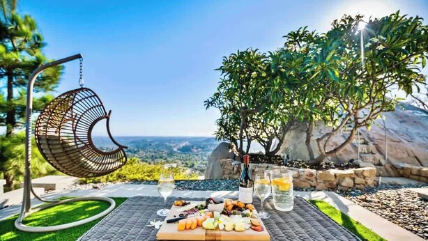 A wicker table with a charcuterie board, a hanging chair, and a bottle of wine, set against a scenic outdoor backdrop.