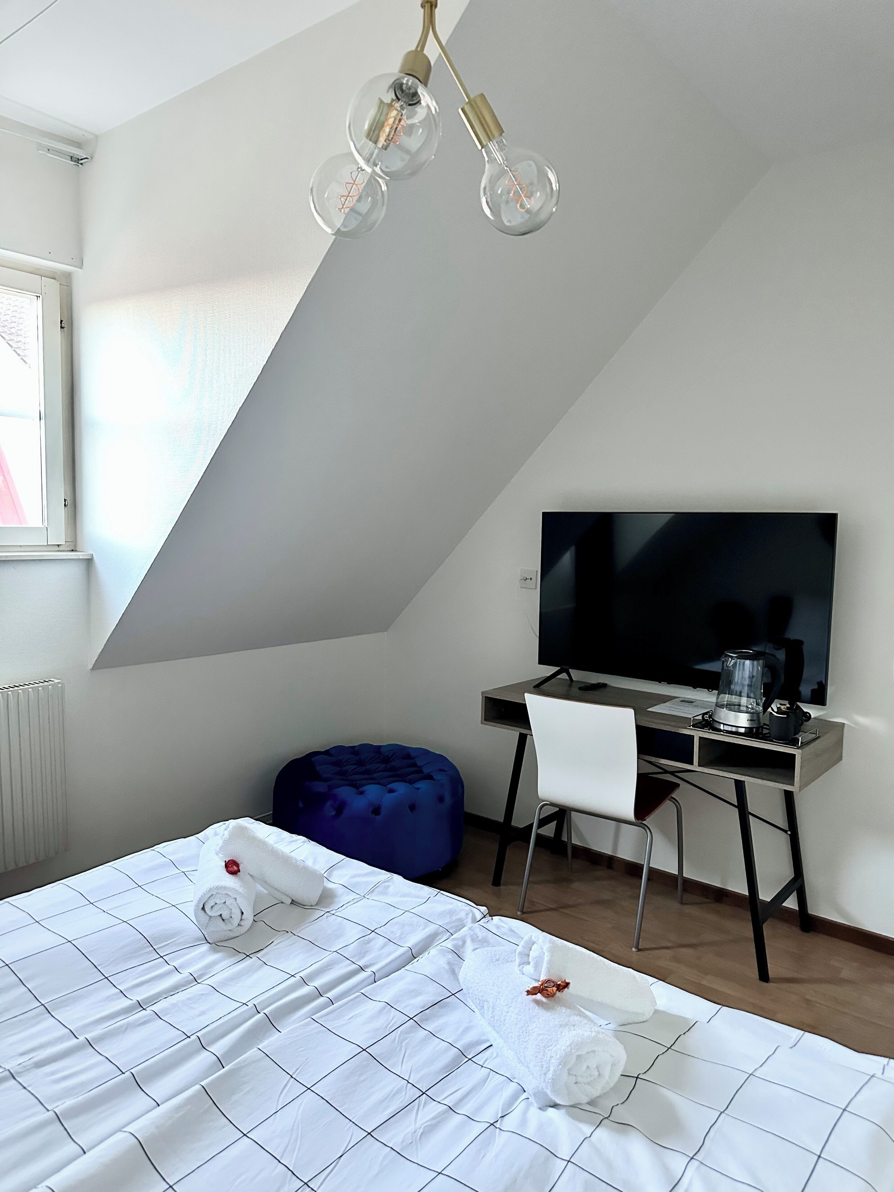 A modern hotel room with a bed, a desk with a computer, a TV, and a unique ceiling light fixture.