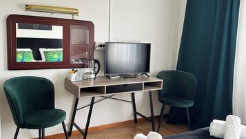 Habitación de hotel con una cama, dos sillas verdes, un escritorio con televisor, un espejo y una ventana con cortinas.